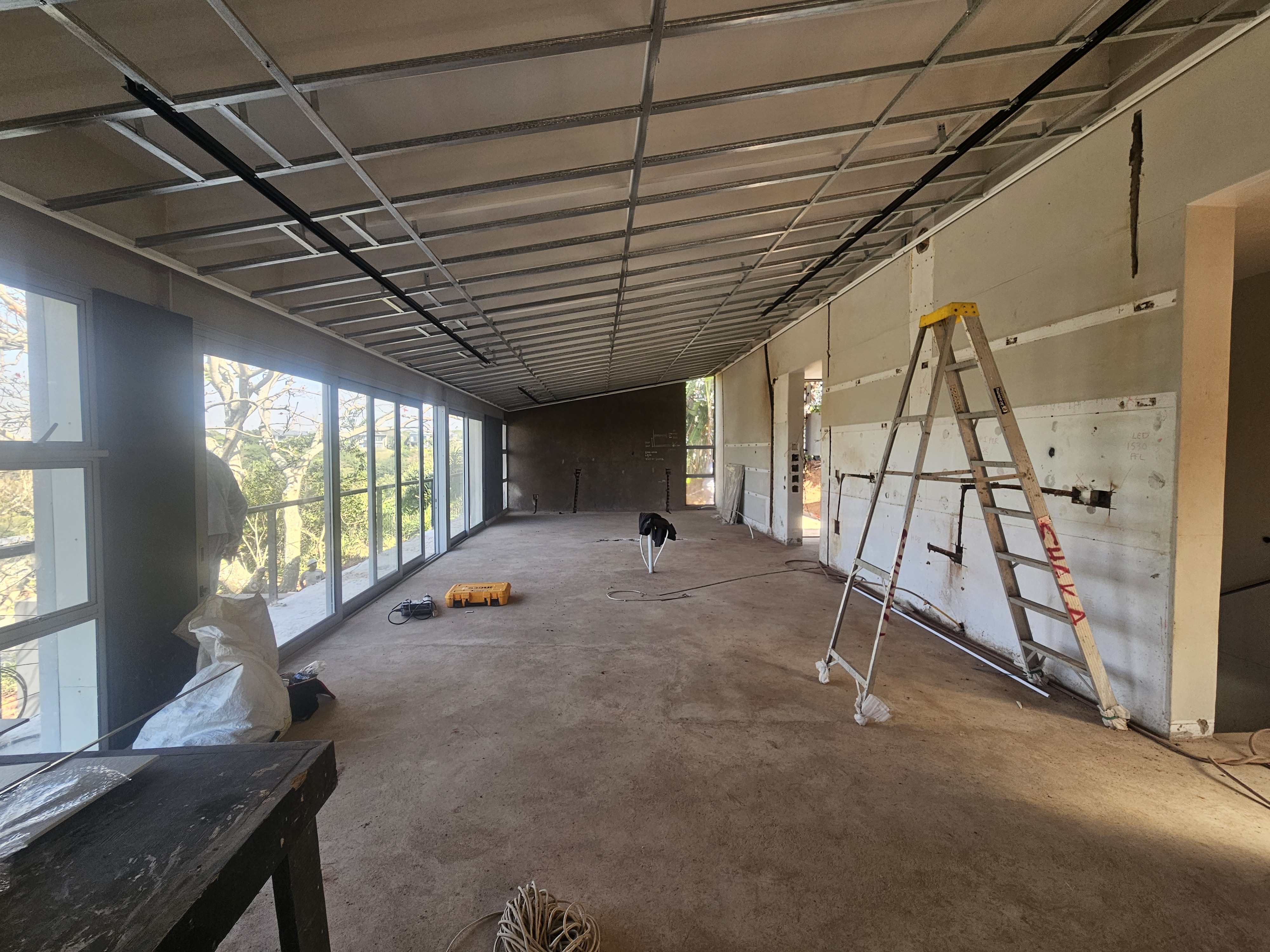 Open plan living room ceiling and drywall construction with full-height windows overlooking lush garden